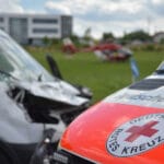Rettungseinsatz mit Krankenwagen und Hubschrauber auf Wiese vor modernem Gebäude im Hintergrund.