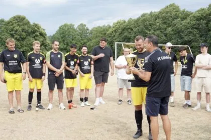 Fußballteam erhält Pokal im Freien bei Siegerehrung, Spieler und Trainer feiern Trophäe auf dem Spielfeld.