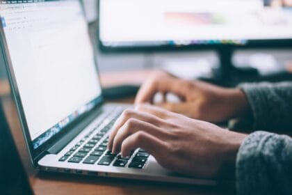 Hände tippen auf Laptop-Tastatur, Arbeiten im Büro oder Homeoffice, produktives Schreiben, Bildschirme im Hintergrund.
